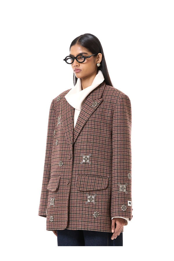 A model wearing a brown and red plaid double-breasted blazer with embroidered details, a white turtleneck, and black glasses, photographed against a solid white background.